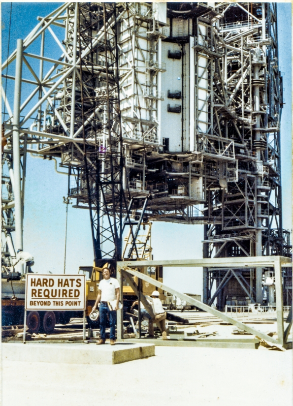 Image 055. Our earliest look at James MacLaren at Space Shuttle Launch Complex 39-B, Kennedy Space Center, Florida, after returning to the jobsite following his layoff from Sheffield Steel, and his subsequent hiring by Ivey Steel. In the far distance, the RSS and FSS loom into the sky, overflowing the frame, sensibly-complete in general appearance, but far from complete for the purposes which they were designed and constructed for, servicing the Space Shuttle, prior to launch. In the near distance, the yellow 90-ton P&H crane is being used to offload more steel from the back of a flatbed semitrailer. Union Ironworkers from Local 808 work the load from on top of the trailer and on the ground next to it, while the white-shirted truck driver lounges against the lopped-off tube steel top of the East Stair Tower. In his left hand, MacLaren grips a clipboard holding the sheets of the shipping list that came with the load of iron, with a yellow notepad behind it. In his right hand, he holds his hard hat, and if you look very close, you can also see a yellow highlighter in that hand for marking up the shipping list indicating items which have been checked and verified as being included in this delivery. On his right hip, a 25-foot tape measure that stayed with him wherever he went on the Pad Deck or on the Towers. Another day on the job. Photo by James MacLaren.